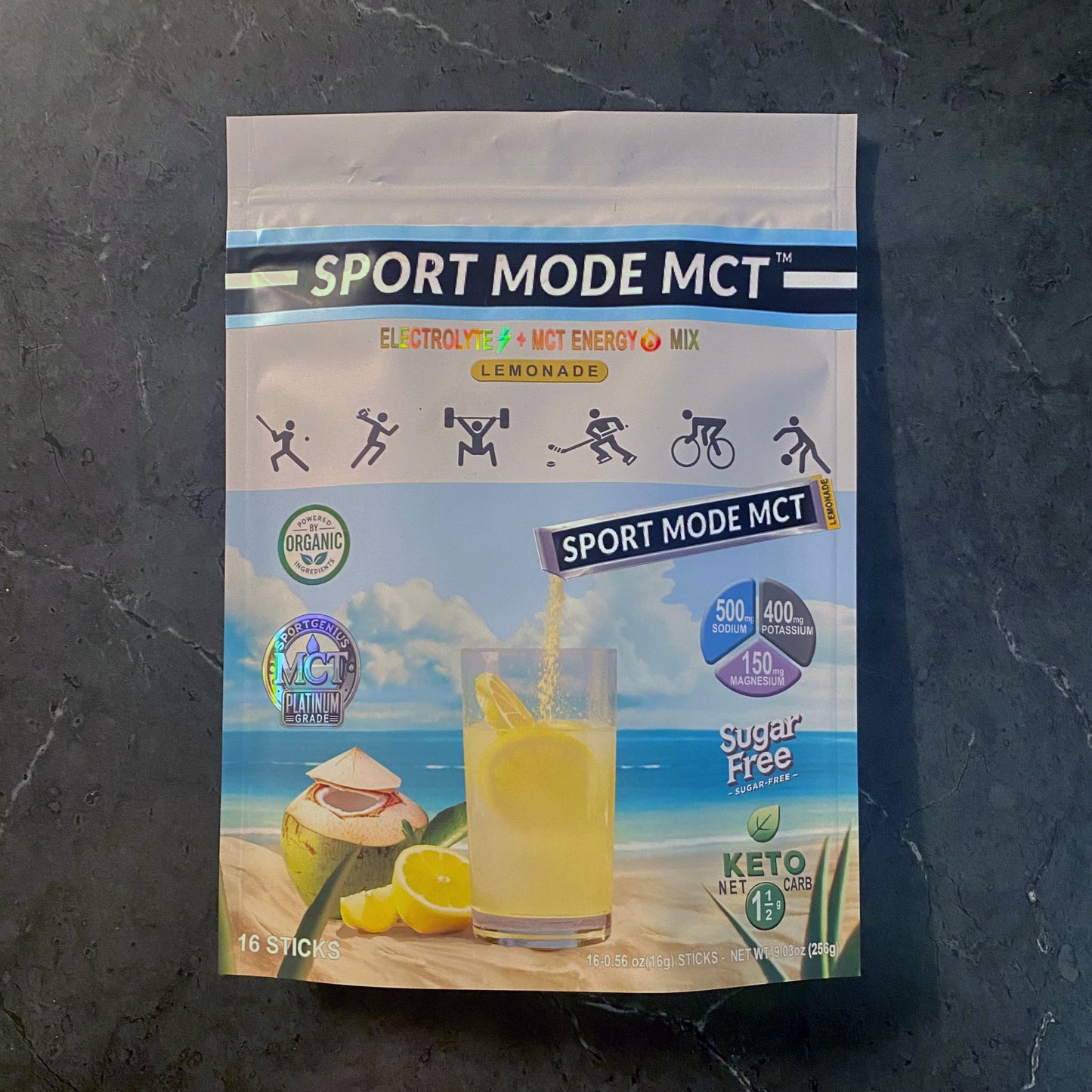 Sport Mode MCT electrolyte and MCT energy drink mix in a resealable lemon-flavored bag shown on a sandy beach theme with ocean and blue sky. The front shows keto-friendly and sugar-free icons, a glass of yellow lemonade, and a lemon drink powder stick being poured into the glass